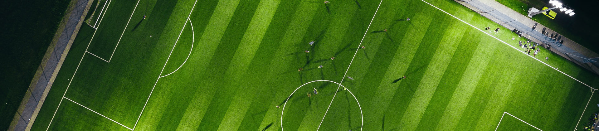 Aerial view of a green soccer field with players scattered around, their shadows cast long on the grass. Spectators and lights are visible along the edge of the field.