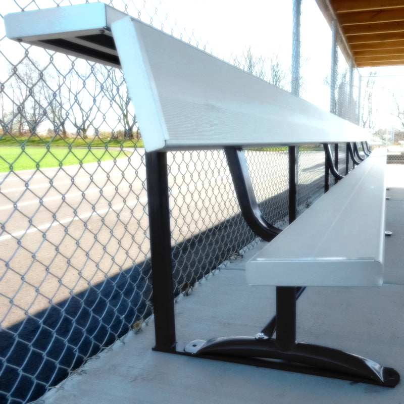 A Team Bench With Storage Shelf, featuring a powder-coated steel frame and shade cover, sits on concrete near a chain-link fence at an outdoor sports field with the empty field visible in the background.