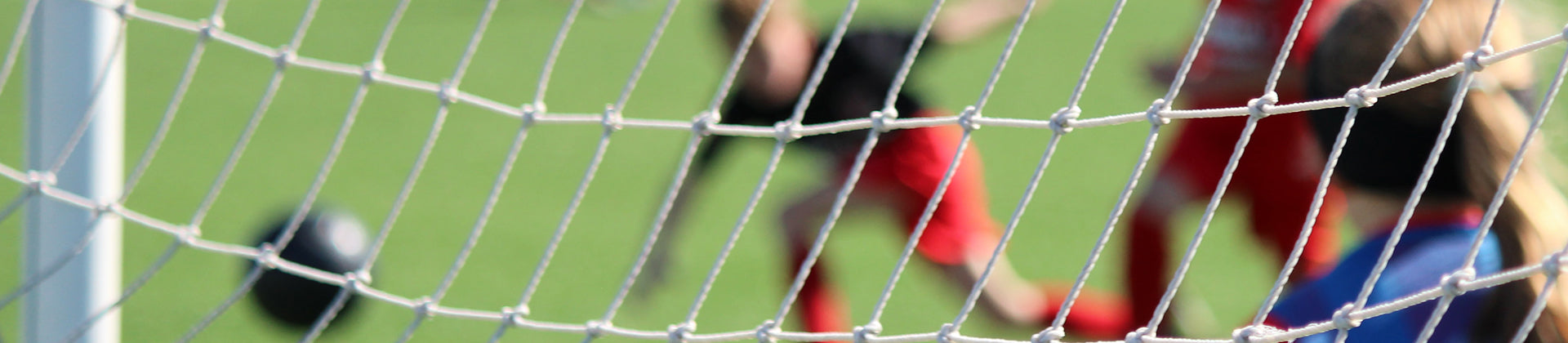 Close-up of a soccer net with blurred players and a soccer ball in the background on a green field, suggesting action near the goal.
