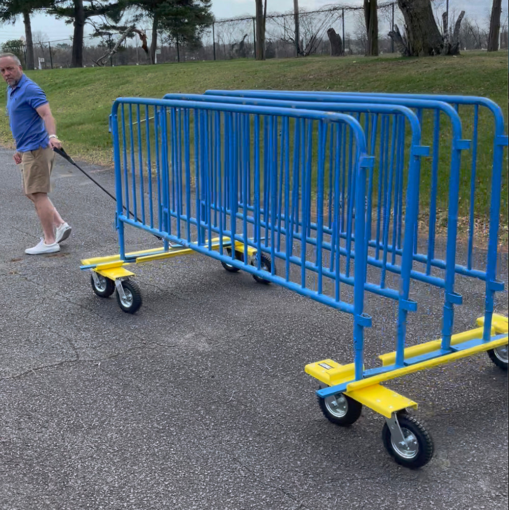 A man in shorts and a blue shirt uses the Goal Taxi to transport blue metal crowd control barriers on an athletic equipment dolly with heavy-duty pneumatic wheels along a paved park-like path.