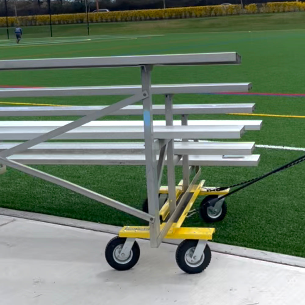 The Goal Taxi, equipped with metal bleachers on heavy-duty pneumatic wheels, is pulled across an artificial turf field, showcasing field markings and efficient goal transportation using this athletic facility equipment dolly.