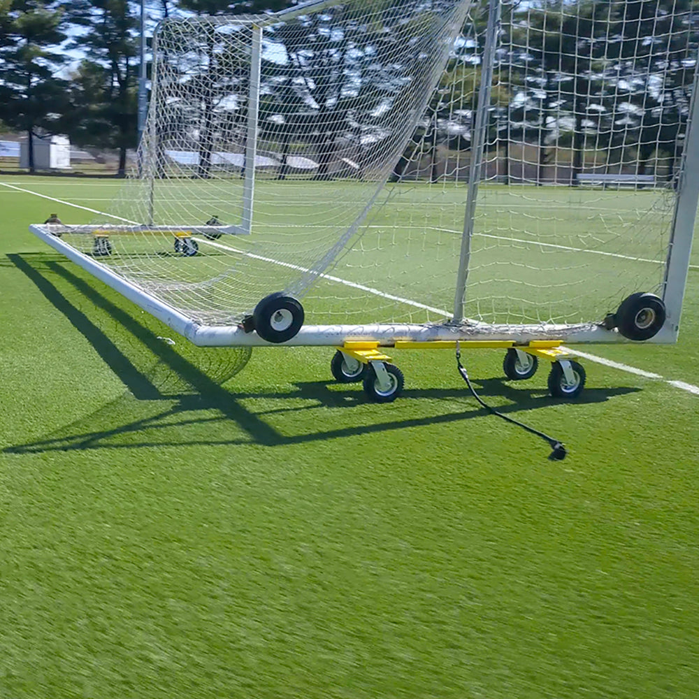 The Goal Taxi, a soccer goal on heavy-duty pneumatic wheels, sits on green artificial turf. Its shadow falls on the field as trees stand in the background under a clear sky, making goal transport simple and efficient.