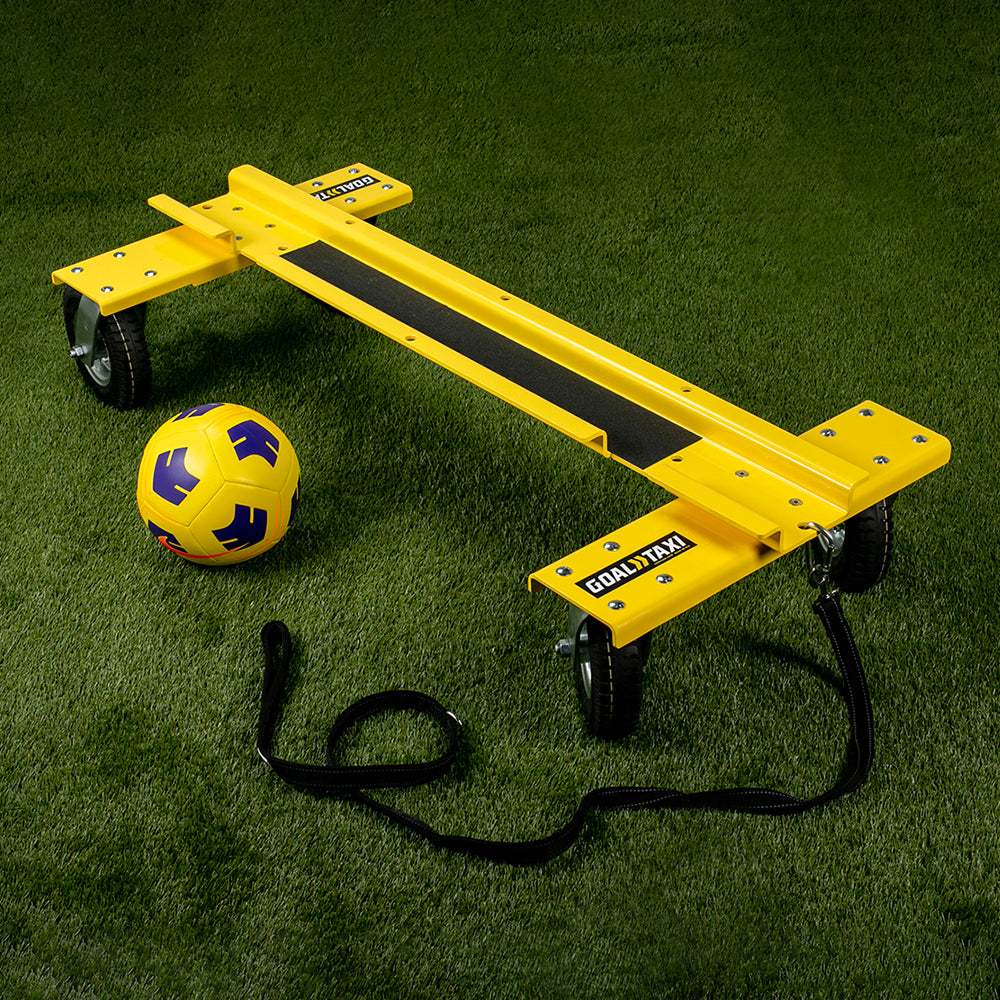 The Goal Taxi, a yellow wheeled tool for transporting goals with heavy-duty pneumatic wheels, is placed on green artificial grass next to a yellow and blue soccer ball and a black strap.