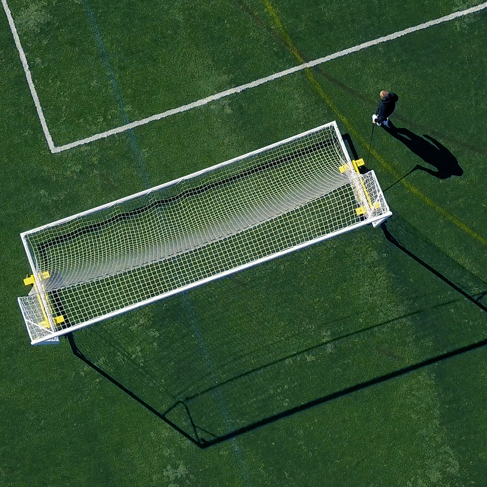 A person stands alone on a green soccer field near a white goalpost, casting a long shadow in the sunlight. A Goal Taxi with heavy-duty pneumatic wheels sits nearby, ready to transport the goal.