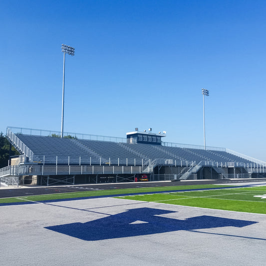 Grandstand Seating with corrosion-resistant aluminum construction and a durable press box overlooks a well-maintained football field under a clear sky, while tall stadium lights rise above the stands.
