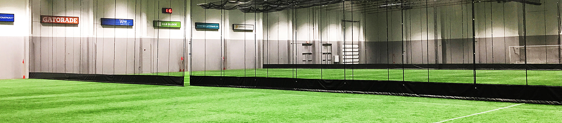 An indoor sports facility with artificial turf fields, separated by black netting. The space is well-lit, and several signs are displayed on the walls above the playing areas.