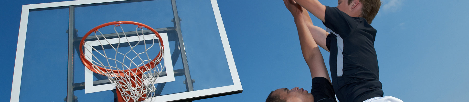 Two people playing basketball outdoors; one player is jumping to block or dunk the ball near the hoop, while the other is closely contesting the play under a clear blue sky.
