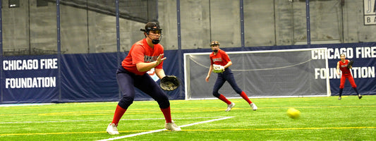 A softball player in a red and navy uniform prepares to catch a rolling ball on artificial turf, while two teammates ready themselves in the background inside an indoor sports facility.