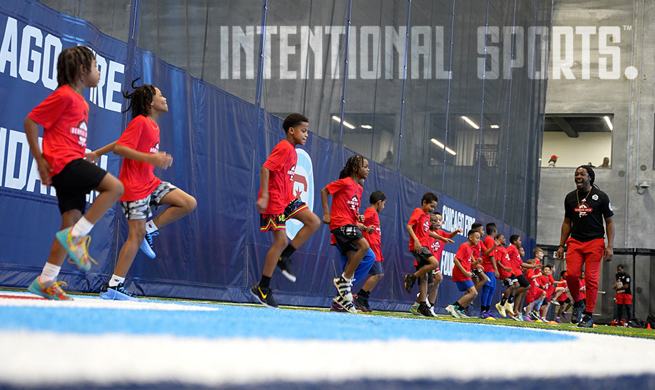 A group of kids in red shirts participate in a sports drill indoors, jumping in a line on artificial turf, led by a coach; INTENTIONAL SPORTS is displayed on the wall behind them.