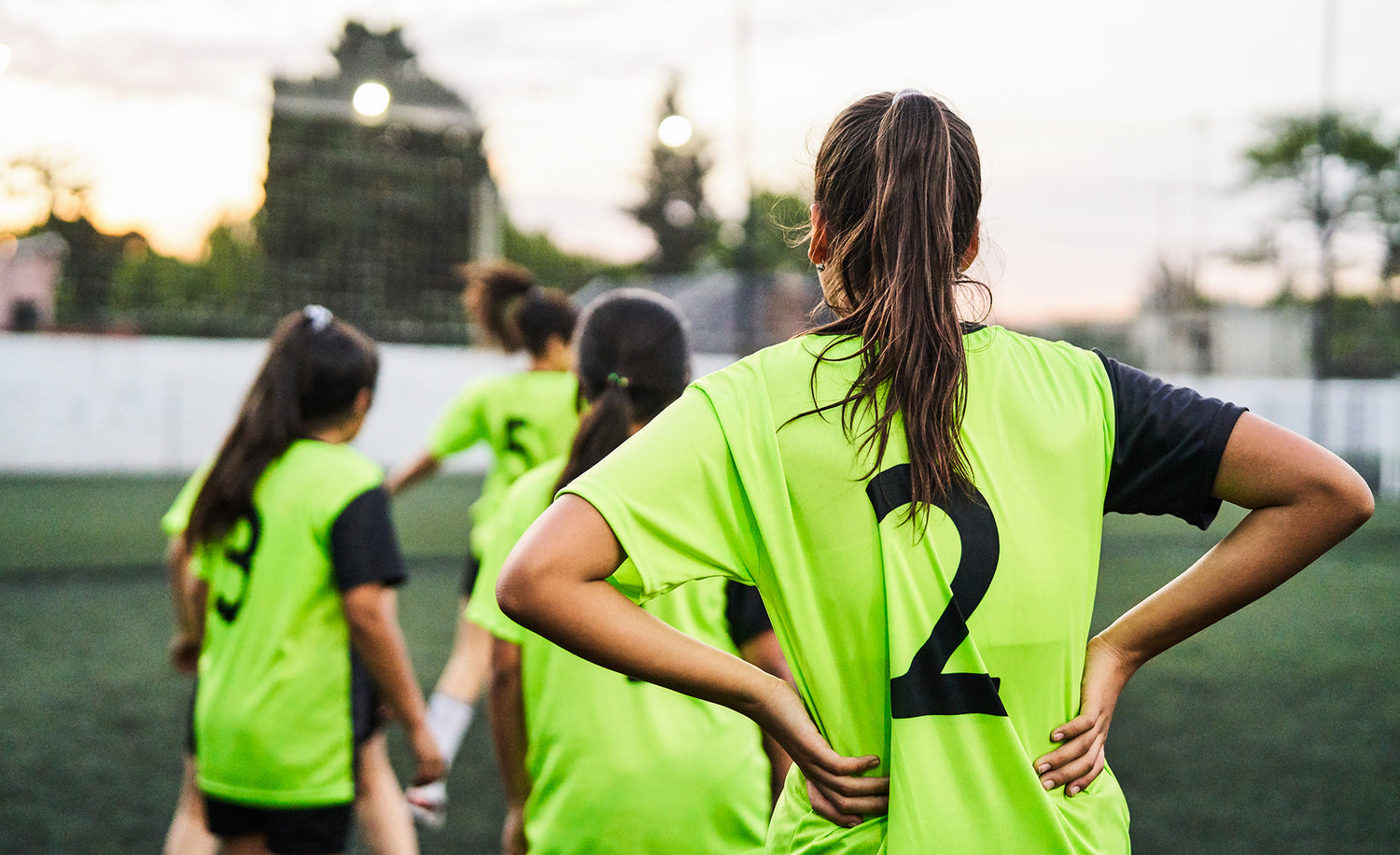 A group of young girls wearing bright green soccer jerseys with black numbers stand on a field, forming a line, with their backs to the camera as they prepare for practice or a game at sunset.