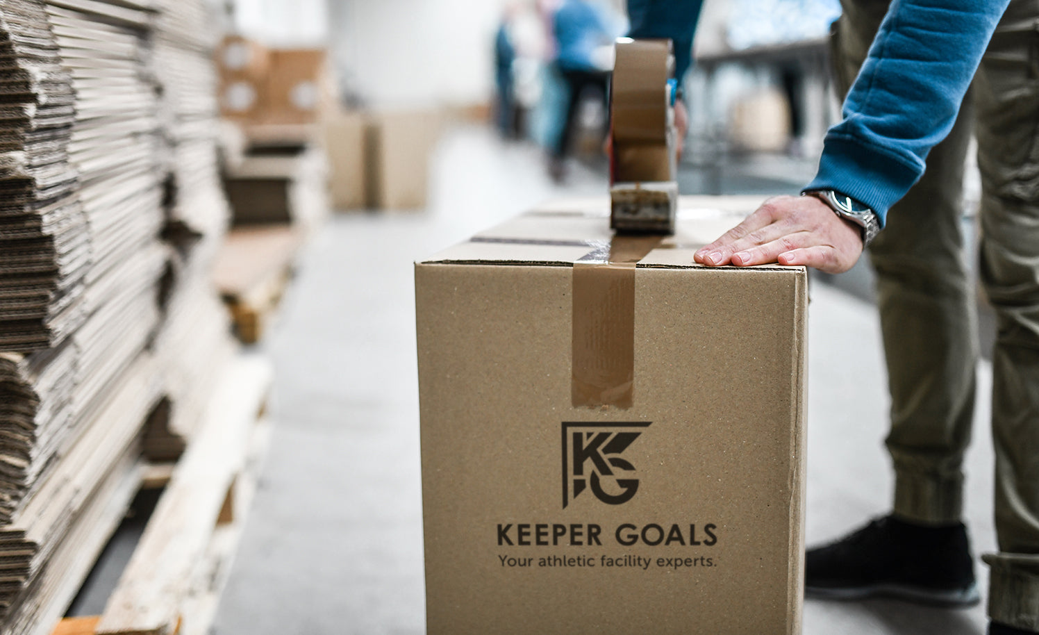 A person seals a cardboard box labeled KEEPER GOALS Your athletic facility experts with tape in a warehouse, surrounded by stacks of boxes and pallets.