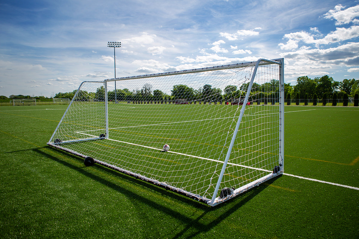 A soccer goal stands on a green artificial turf field under a partly cloudy sky, with a soccer ball resting near the center of the goal. Trees and stadium lights are visible in the background.