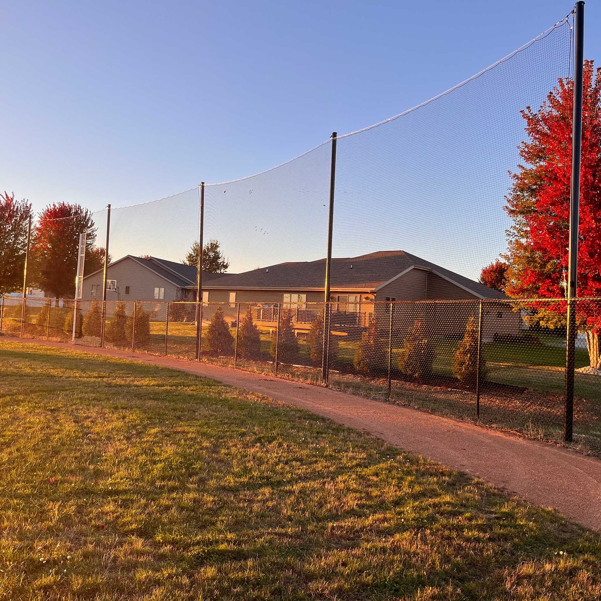 A paved path runs beside Standard Baseball and Softball Backup Netting Systems and a chain-link fence, with houses and trees featuring red autumn leaves visible in the golden hour background.