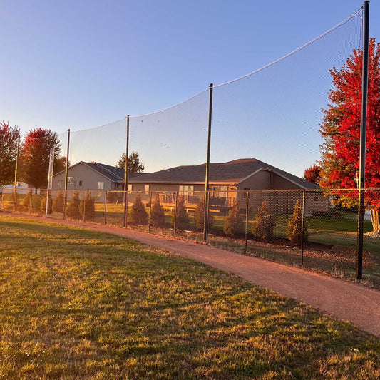 A paved path runs beside Standard Baseball and Softball Backup Netting Systems and a chain-link fence, with houses and trees featuring red autumn leaves visible in the golden hour background.