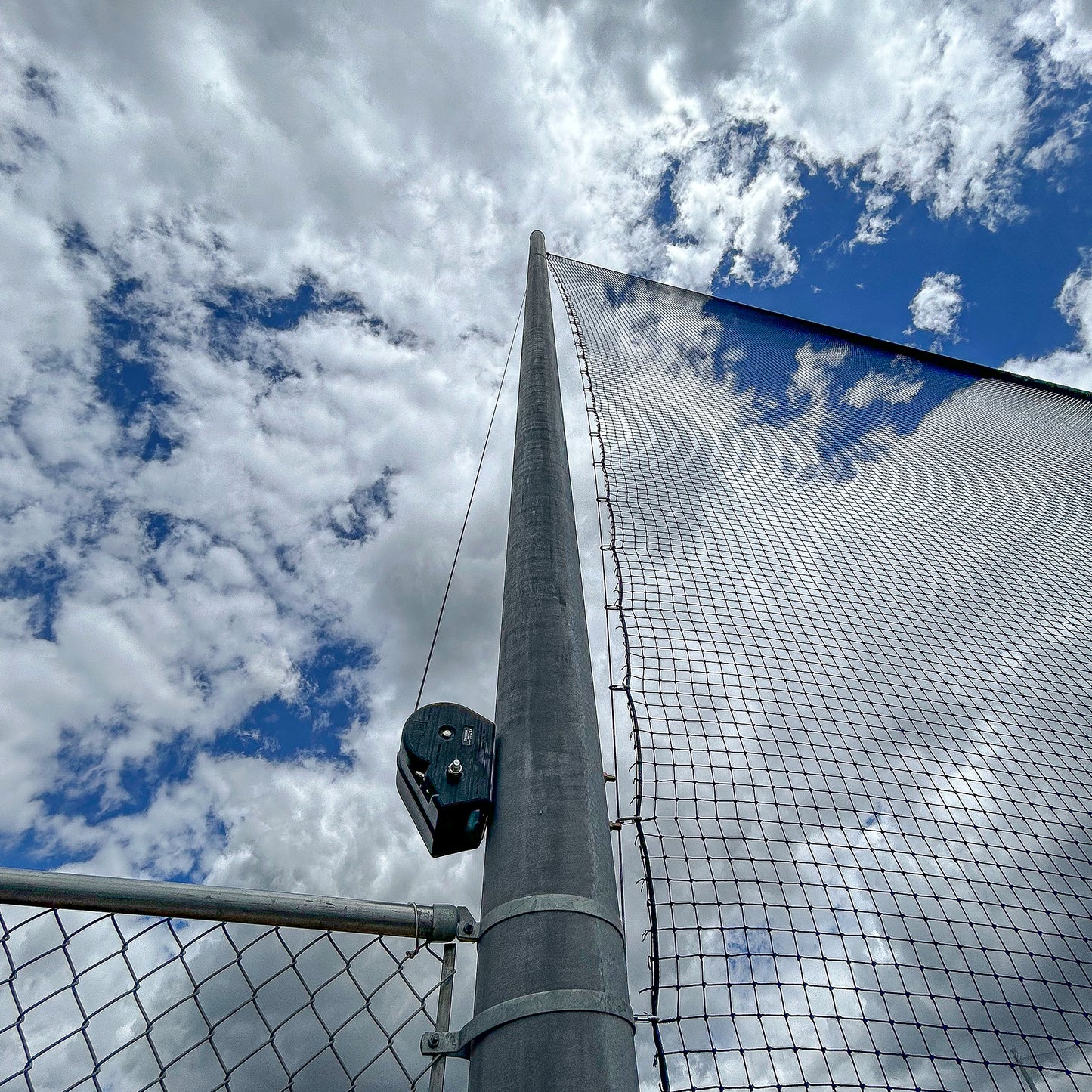 A tall metal pole supports Custom Engineered Backstop and Backup Netting Systems, with dramatic clouds and patches of blue sky in the background, viewed from below at an upward angle.