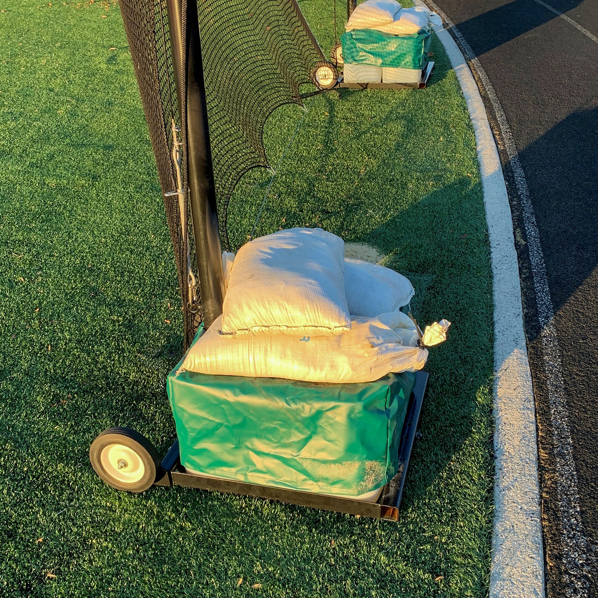 The Portable Lacrosse Backup Netting System, featuring durable mesh and a green padded base, stands on artificial turf beside a curved track, ready for high-impact play under bright sunlight.