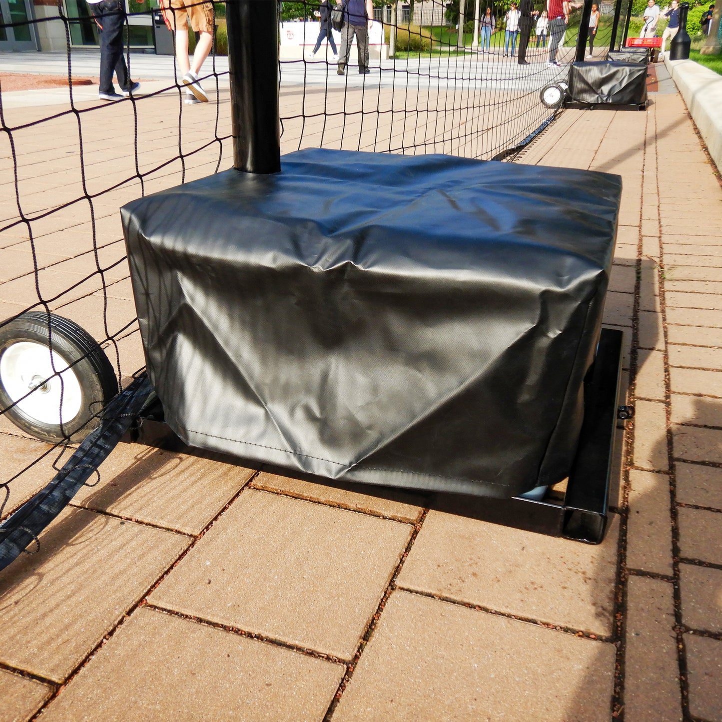 Close-up of the Portable Soccer Backup Netting System’s black wheeled base on pavement, supporting durable netting outdoors, with people walking and standing in the background.
