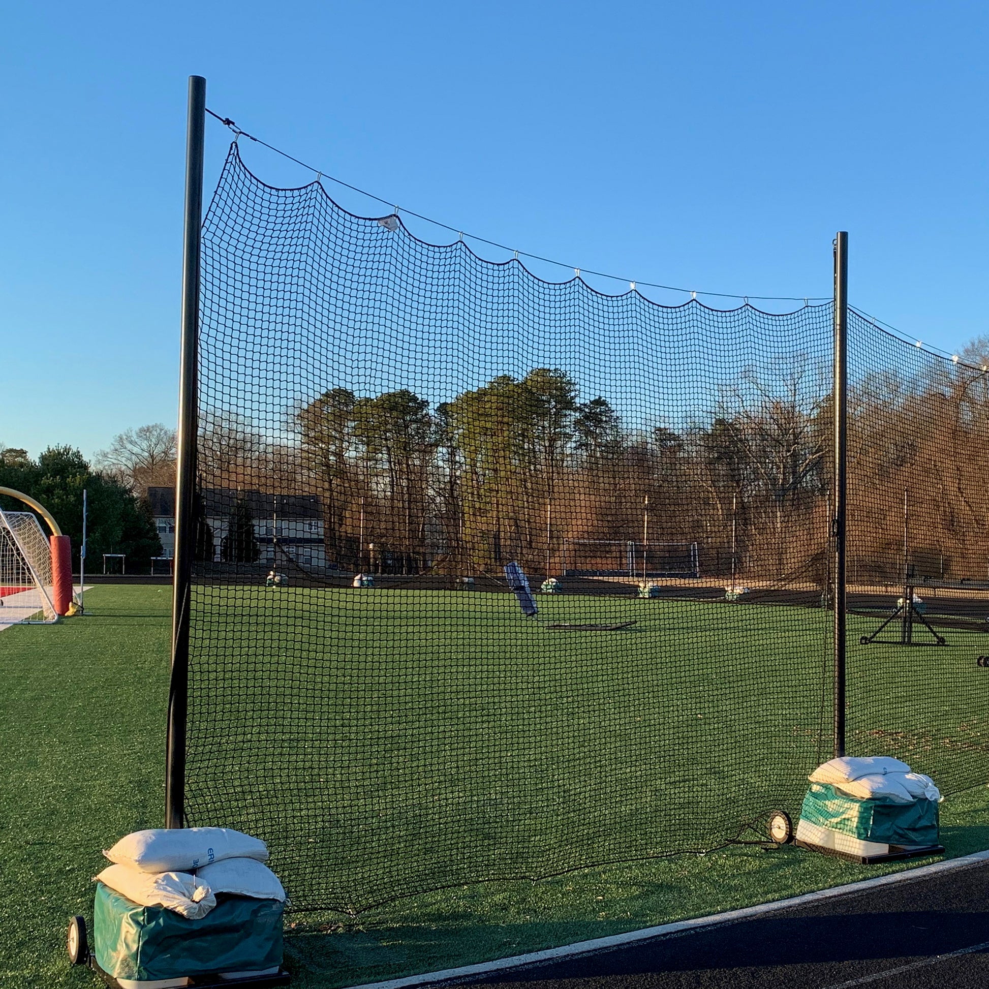 A Portable Lacrosse Backup Netting System with durable black mesh is set up on a green sports field, anchored by sandbags. Trees and buildings border the empty field beneath a clear blue sky.