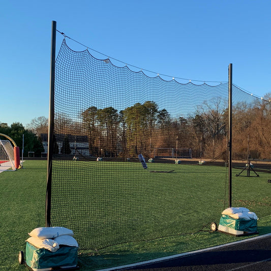 A Portable Lacrosse Backup Netting System with durable black mesh is set up on a green sports field, anchored by sandbags. Trees and buildings border the empty field beneath a clear blue sky.