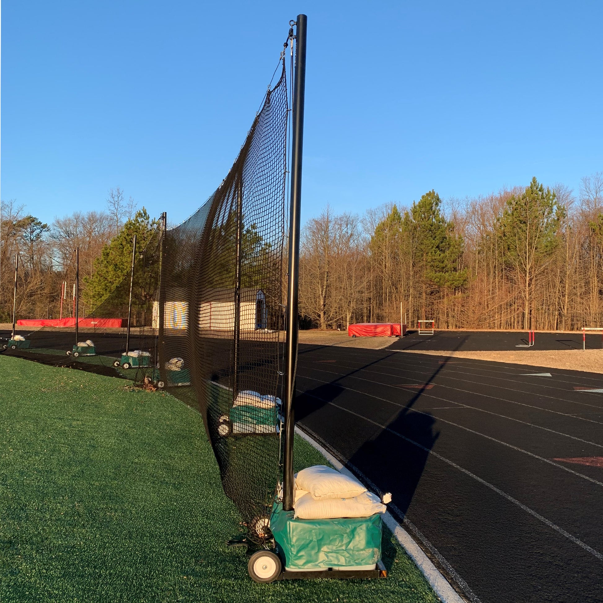 The Portable Lacrosse Backup Netting System, featuring durable mesh and wheeled support poles, stands along the edge of a green artificial turf field by a running track, ready for high-impact play under a clear blue sky.