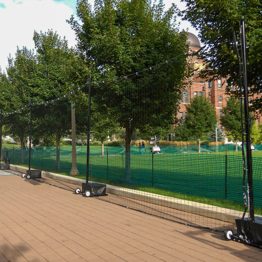 The Portable Soccer Backup Netting System, with temporary black mesh on wheeled bases, lines a paved walkway next to a grassy park, mature trees, and a large brick building in the background.