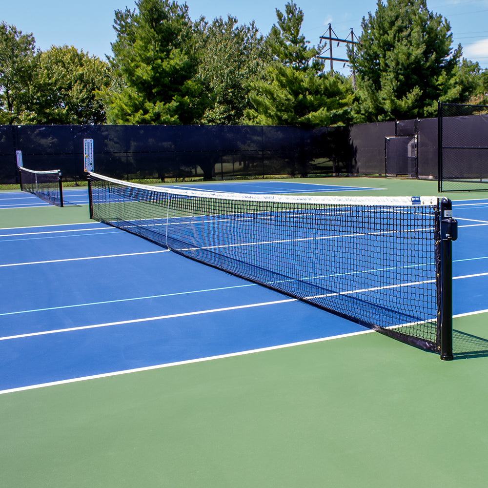A blue and green outdoor tennis court with a black net supported by DTP Heavy-duty 3 OD Tennis Posts, surrounded by trees and a black fence under a sunny sky.