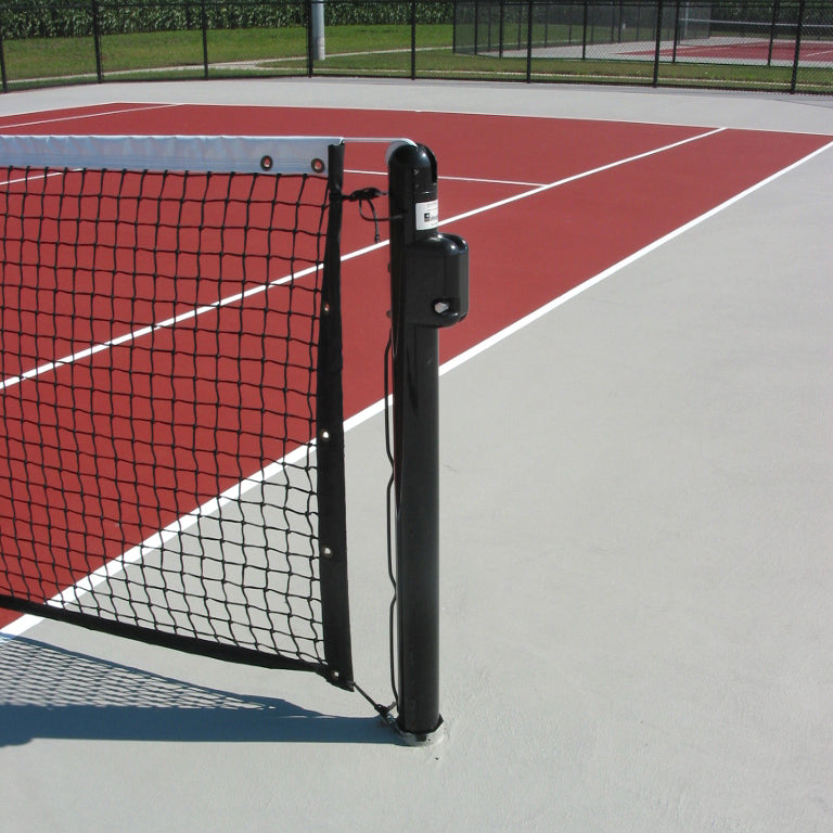 A close-up of DTP Heavy-duty 3 OD Tennis Posts and a black net with welded lacing rods on a red and gray outdoor tennis court, surrounded by a fence. The court is empty.
