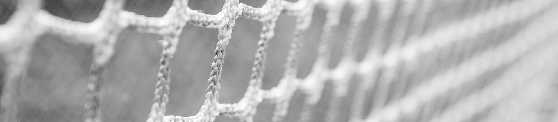 Close-up, angled view of a woven rope net or mesh, shown in black and white. The image focuses on the texture and pattern of the intersecting ropes, with a blurred background.