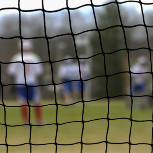 A close-up of Standard Lacrosse Backup Netting Systems with three blurred athletes in blue and white uniforms standing on a grassy field in the background.