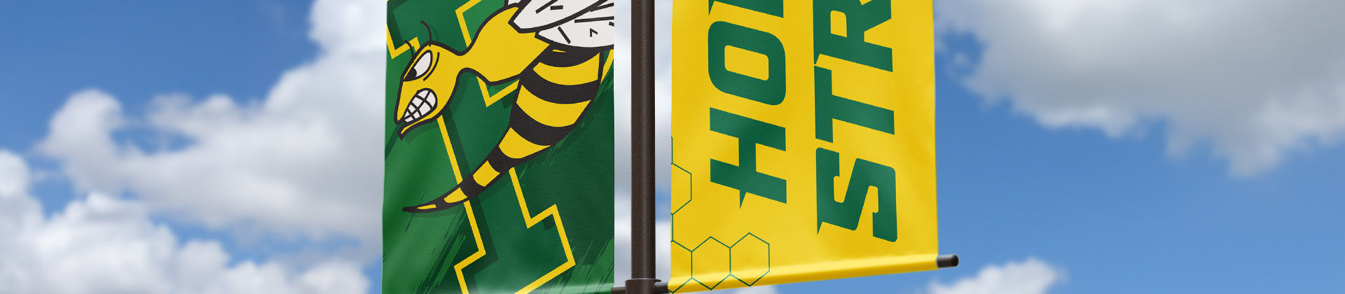 Two vertical banners on a pole; one shows a fierce yellow and black hornet on a green background, the other is yellow with bold green text reading HORNET STRONG, against a blue sky with clouds.