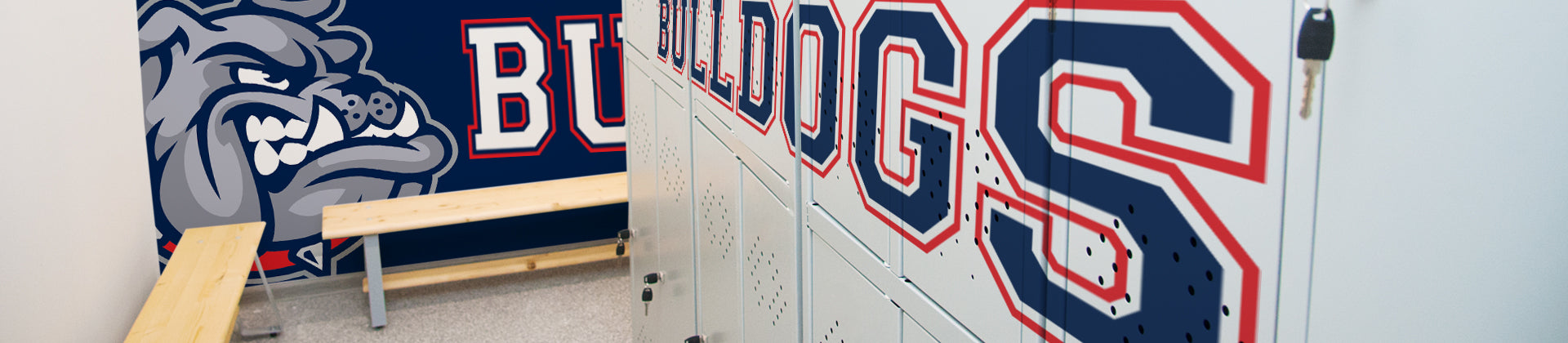 A locker room with white lockers displaying large red and blue letters, wooden benches, and a wall mural featuring a fierce bulldog mascot and the word BULLDOGS in bold, sporty font.