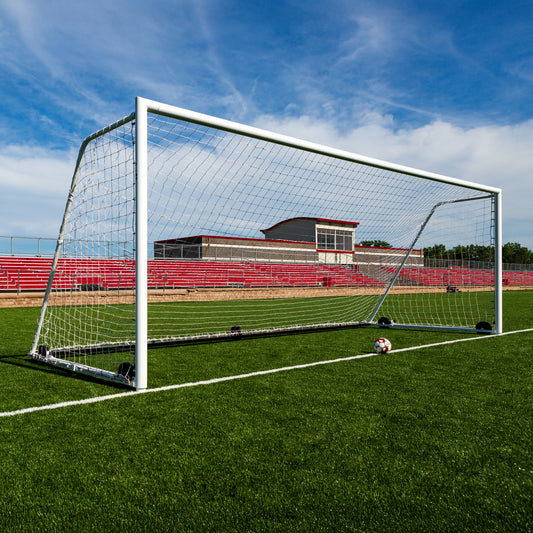 The Ultimate Wheeled Premier Soccer Goals stand on a green field with a ball in front, red bleachers, and a school building in the background beneath a partly cloudy blue sky.