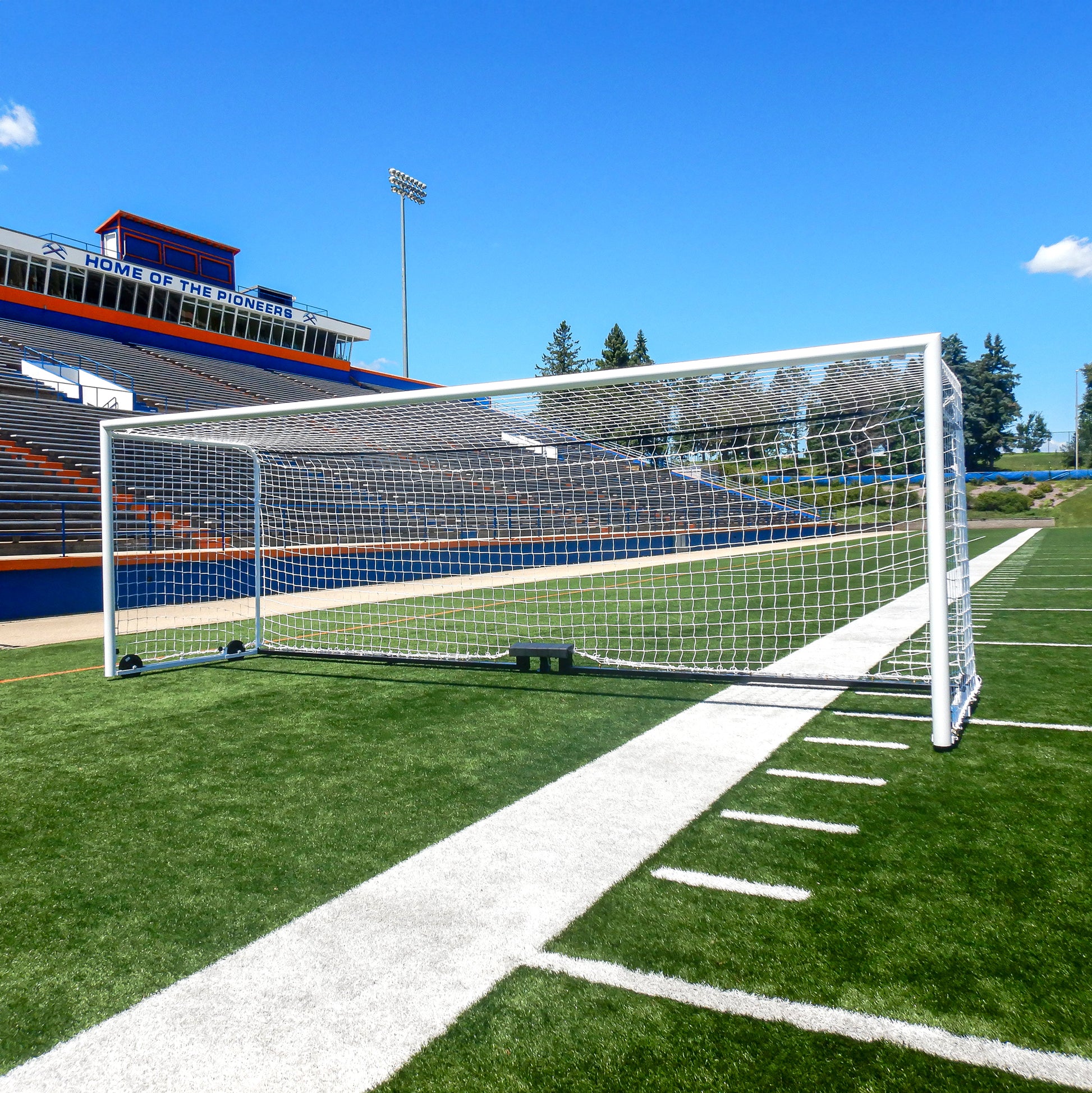 An Ultimate Wheeled Box-Style Soccer Goal stands on a green field with white lines, empty bleachers, and a stadium building labeled Home of the Pioneers visible in the background beneath a blue sky.