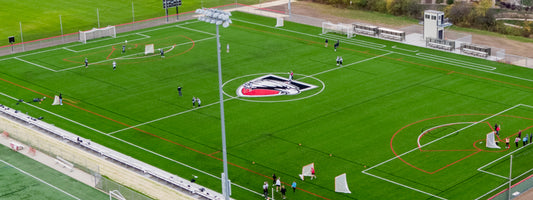 Aerial view of a green sports field with white and red lines, a large bird logo at the center, and groups of people playing and practicing on different parts of the turf. Benches and a small building are along one side.