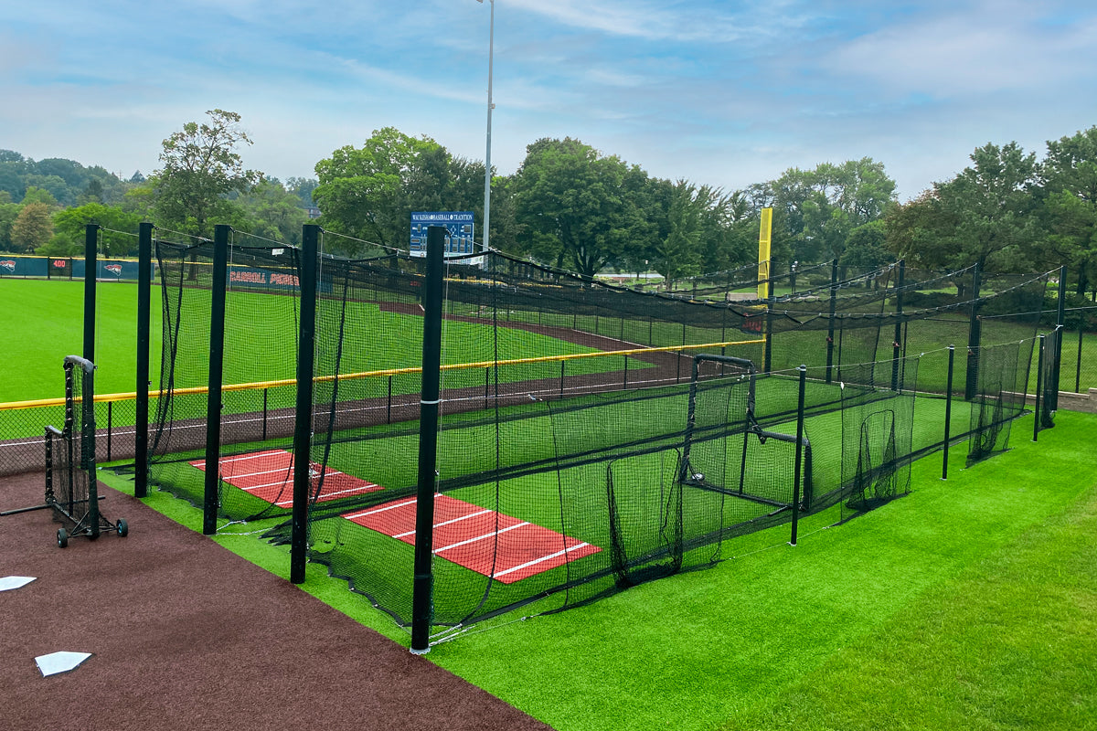 A baseball field with a fenced batting cage featuring two red turf pitching mounds, surrounded by green grass and trees under a blue sky.