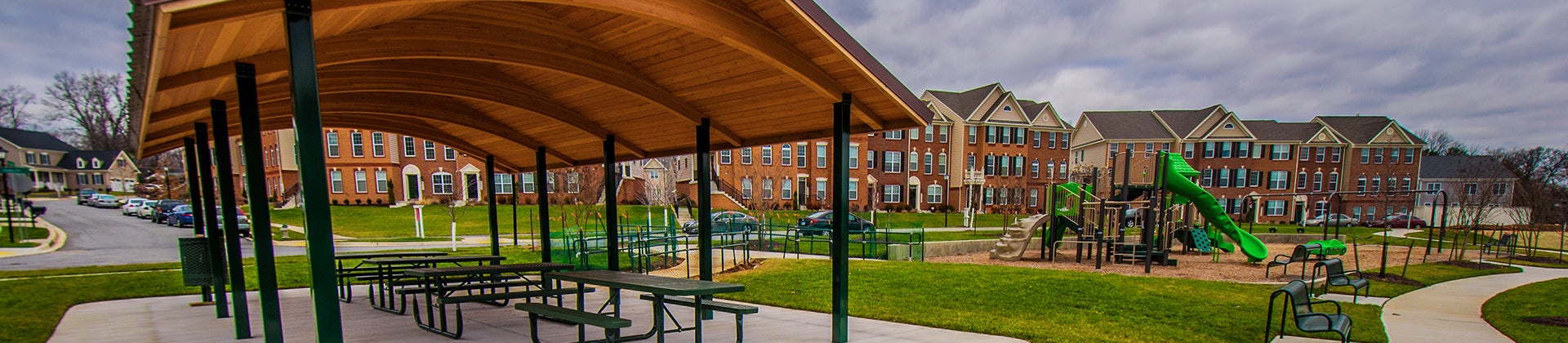 A covered pavilion with picnic tables stands next to a playground with green slides in a grassy neighborhood park, surrounded by rows of brown townhouses under a cloudy sky.