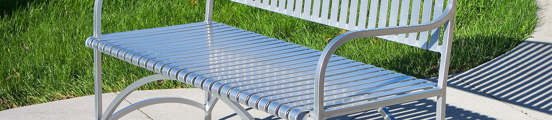 A silver metal bench with slatted seat and backrest sits on a curved concrete pathway next to green grass, casting shadows on the ground.