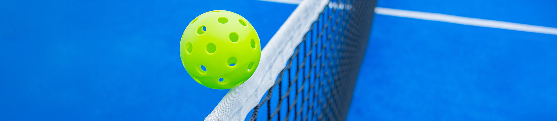 A bright green pickleball hovers above the net on a blue pickleball court, captured in mid-air during play.