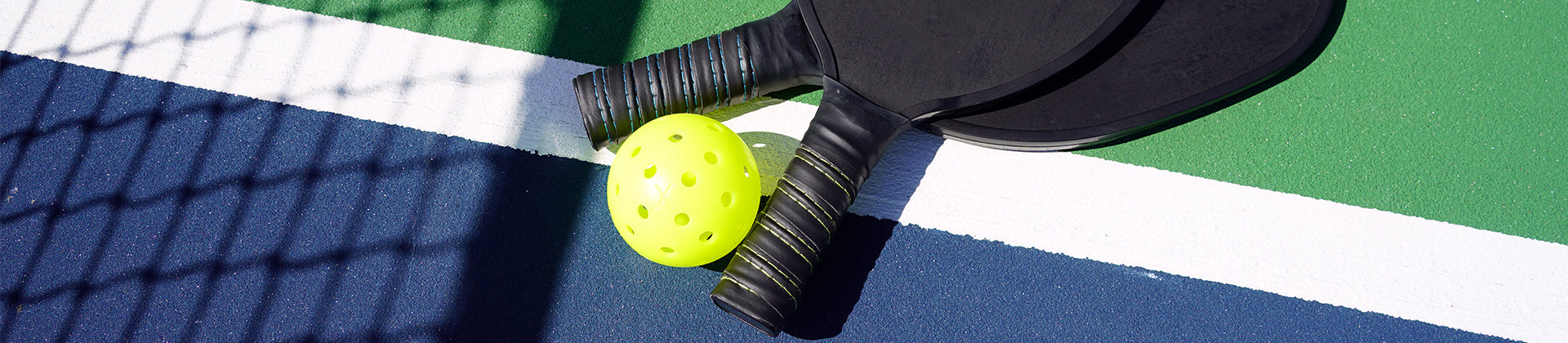 Two black pickleball paddles and a bright yellow pickleball rest on a blue and green court near a white line, with a shadow from a fence visible on the surface.