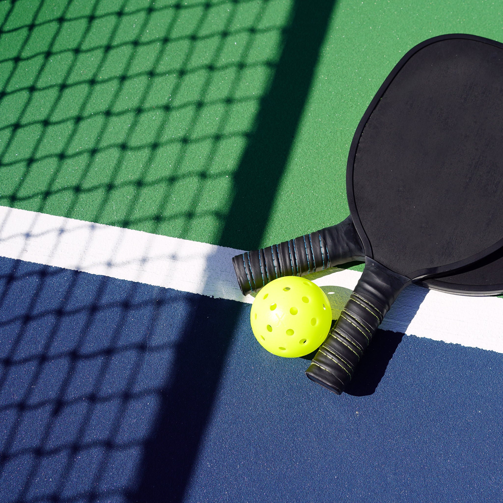 Two black pickleball paddles and a yellow pickleball rest on a court with blue, green, and white lines. A shadow from a chain-link fence falls across the scene.