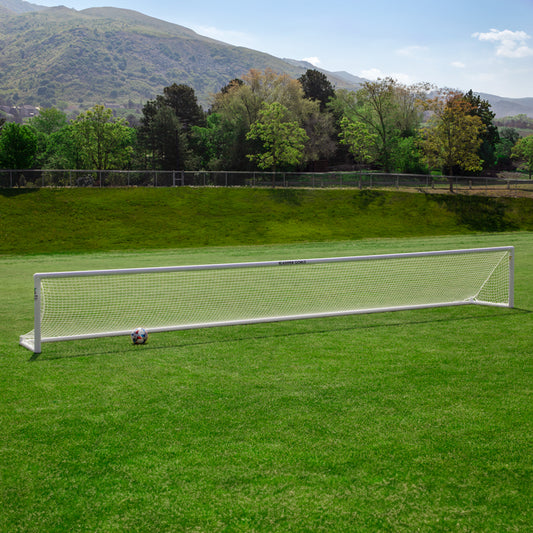 A Precizion Soccer Goal with a soccer ball by one post stands on a green field, ideal for accuracy training, with trees and hills in the background beneath a partly cloudy sky.