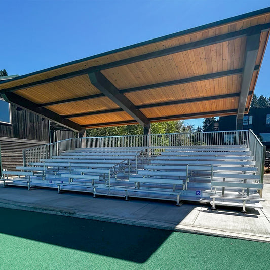 The Premium Hard Shell Bleacher Cover provides shaded metal seating and railings on a concrete base. Its permanent roof offers generous shade, while sunlight fills the area, with blue sky and trees visible in the background.