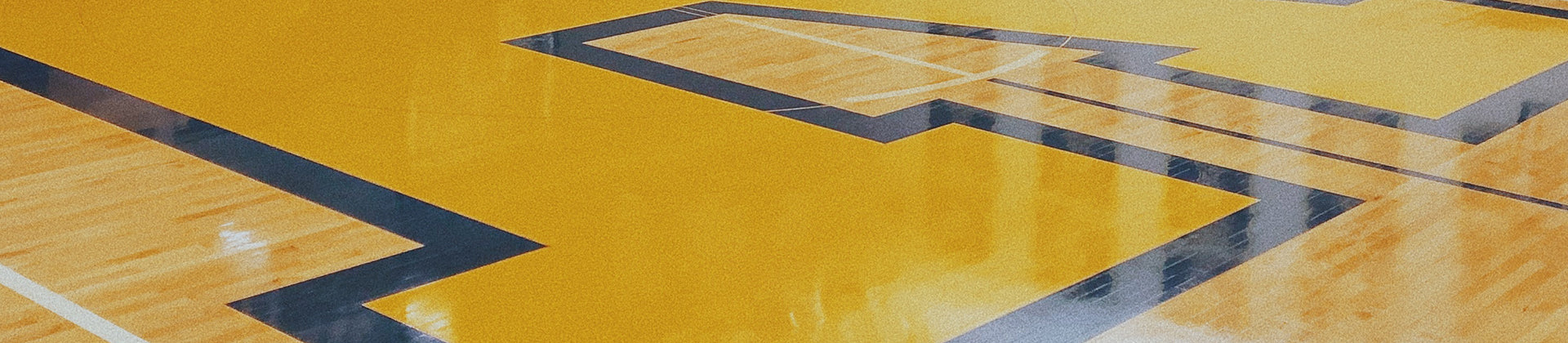 Close-up of a polished gymnasium floor featuring a large yellow area bordered and detailed with dark blue lines, likely forming part of a sports court or a team logo.