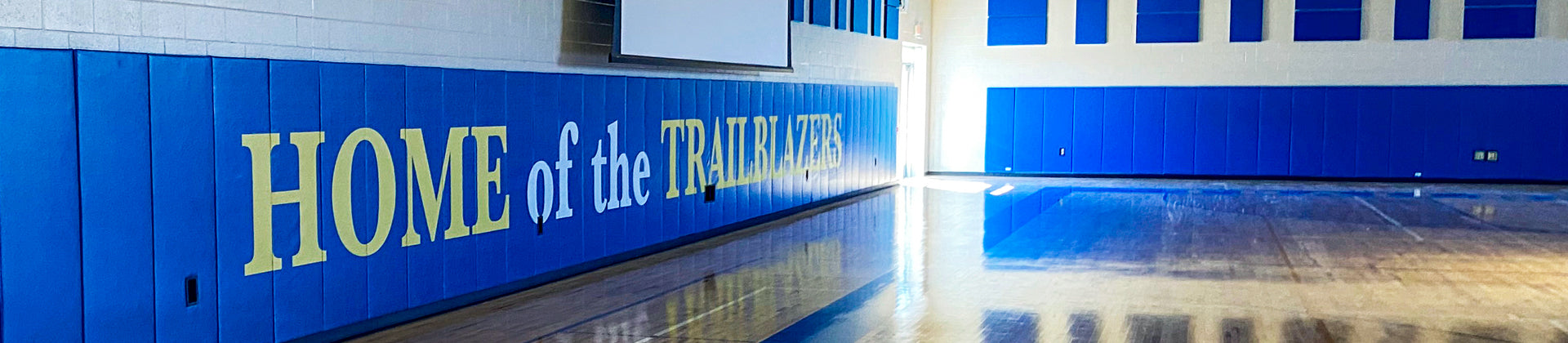 A gymnasium with blue and yellow walls, featuring large text that reads HOME of the TRAILBLAZERS. Sunlight reflects off the polished wooden floor.