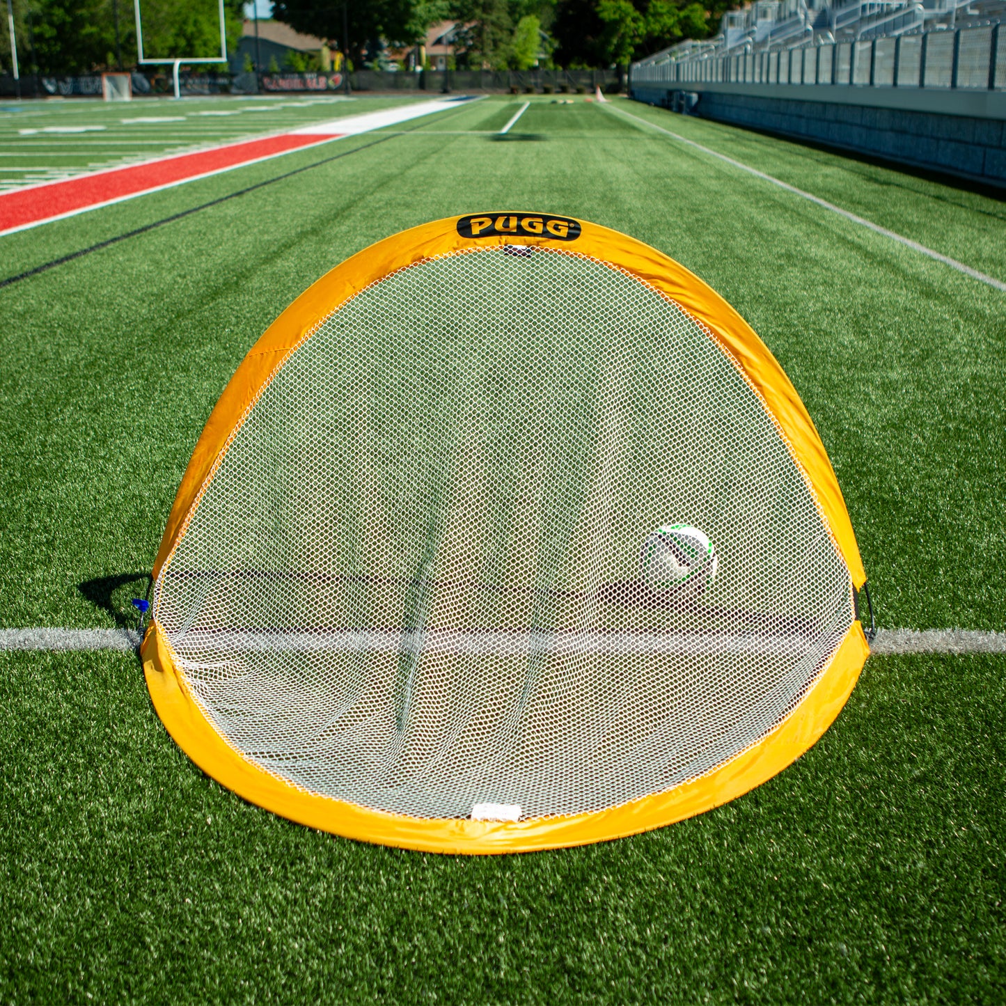 A Pugg Soccer Goal in yellow is set up on a green turf field near a running track, with bleachers and trees visible in the background.