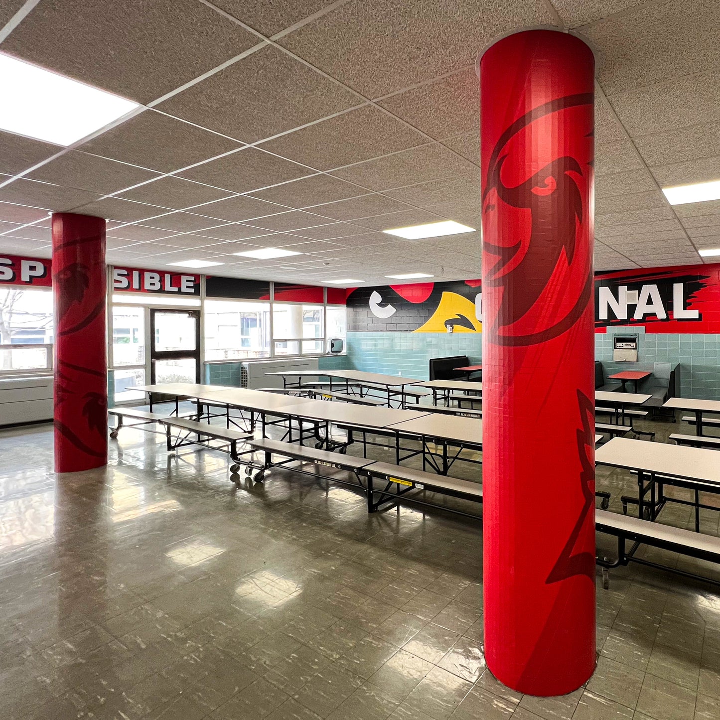 A school cafeteria features long tables, benches, bright lighting, and windows. Large red Pillar Wraps with cartoon bird faces and colorful wall murals with bold text and bird graphics decorate the space.