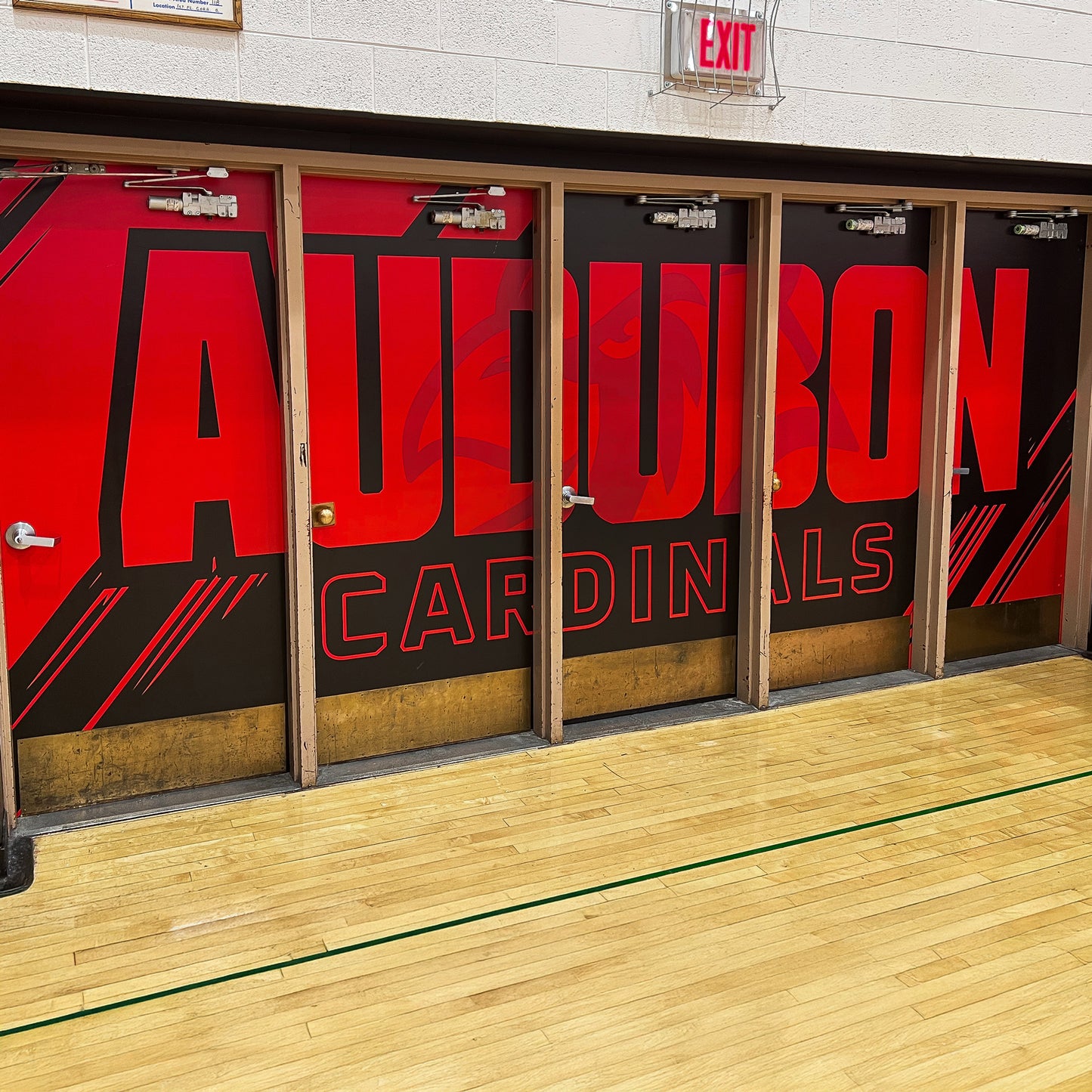 Four gymnasium doors are covered with Custom Vinyl Door Wraps featuring a bold red and black Audubon Cardinals design and a subtle cardinal logo. These wraps boost school branding, with a green line visible on the wooden floor in front.