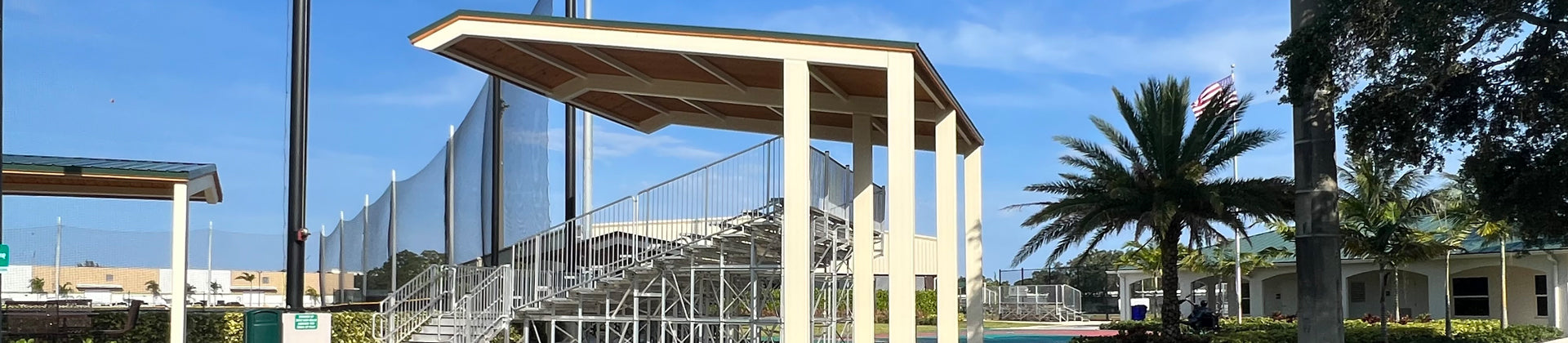 A covered outdoor metal bleacher structure stands beside a sports field, with palm trees, buildings, and a clear blue sky in the background.