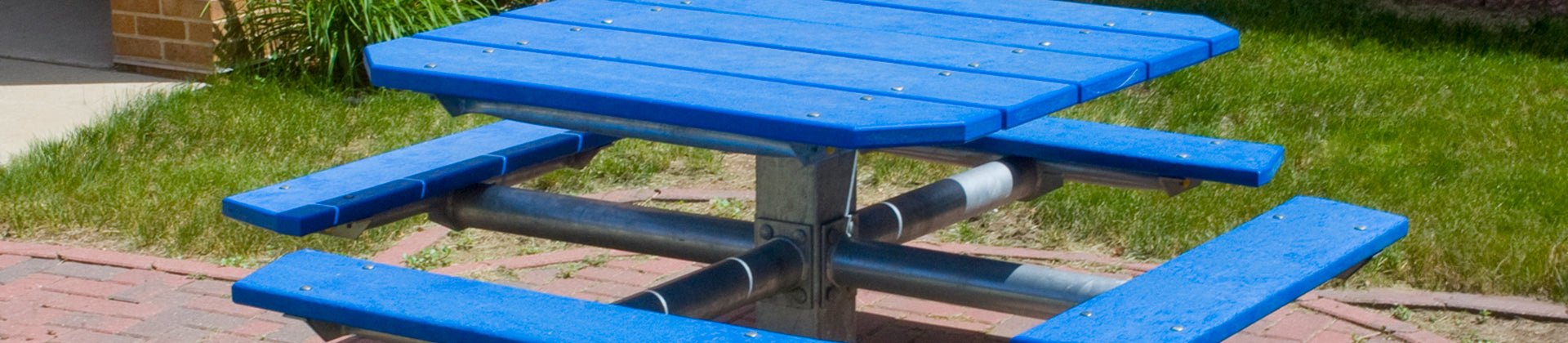 A blue outdoor picnic table with four attached benches sits on a brick patio, surrounded by green grass and some plants near a building corner.