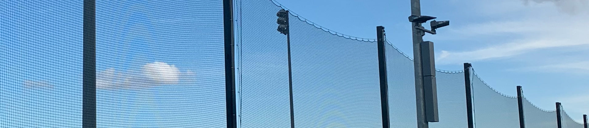 Tall metal poles support a high net fence with lights and a security camera, set against a blue sky with a few clouds.